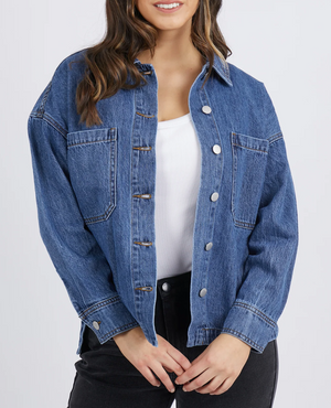 A person stands against a plain, light background with loose, wavy hair. They wear the Foxwood Joelle Denim Shacket, a relaxed fit 100% cotton blue denim piece with front pockets and metal buttons, layered over a white shirt and paired with black pants.
