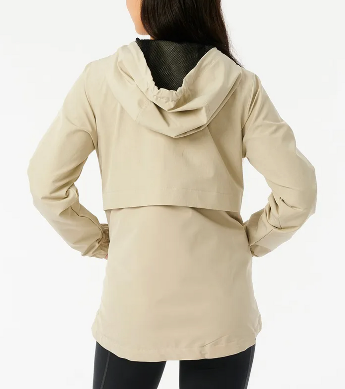 Rear view of a person in a beige Rip Curl Anti Series Elite III Jacket with hands on hips. The water-repellent jacket, featuring mesh lining, adjustable hood, and back flap detail, is paired with black pants against a plain white background.