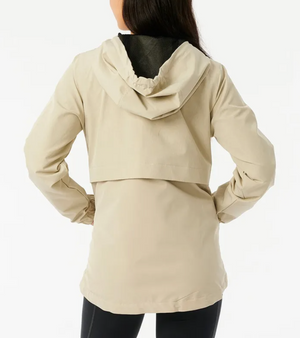 Rear view of a person in a beige Rip Curl Anti Series Elite III Jacket with hands on hips. The water-repellent jacket, featuring mesh lining, adjustable hood, and back flap detail, is paired with black pants against a plain white background.