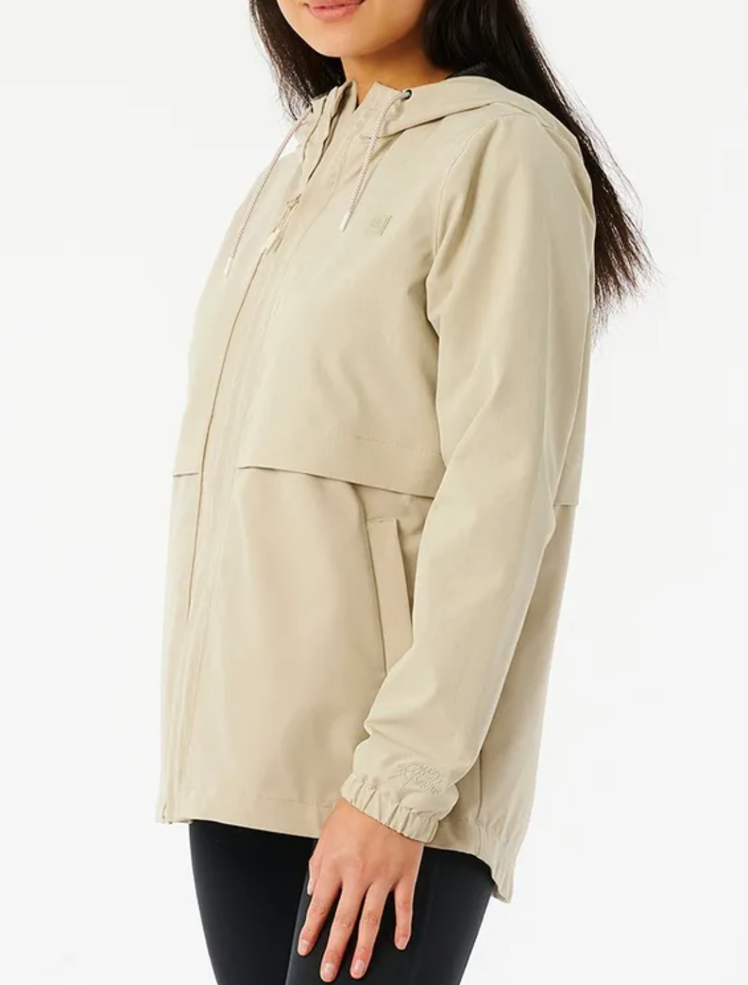 A person wearing a light beige Rip Curl Anti Series Elite III Jacket with long sleeves, an adjustable hood, and a zip-up front stands against a white background. The jacket's drawstrings complement the person's long dark hair as they pair it with black pants.