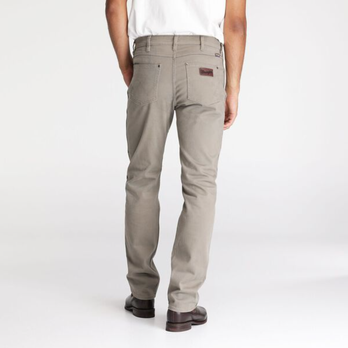 A person is shown from the back wearing Wrangler Classics Straight Moleskin Jeans in gray khaki, featuring visible seams and a leather patch on the back pocket. They are paired with a white shirt in a plain, light-colored studio setting that highlights their water-saving technology.