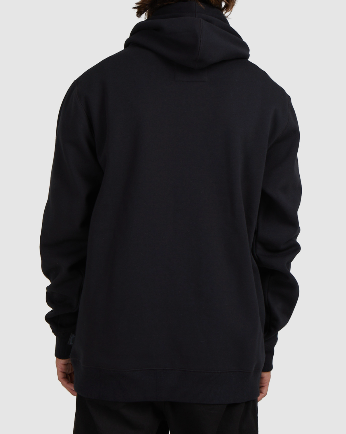 A person is seen from the back wearing a black Billabong Shoreline Pullover with the hood down, standing against a plain light gray background. Their hands rest by their sides, highlighting the pullover’s casual heavyweight style.