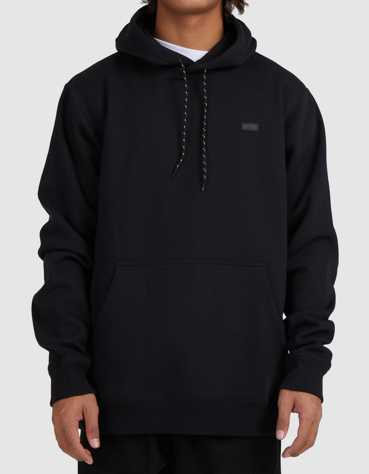 A person wearing a Billabong Shoreline Pullover—a casual black hoodie with a front pocket, black and white drawstrings, and a small rectangular chest patch—stands against a light gray background.