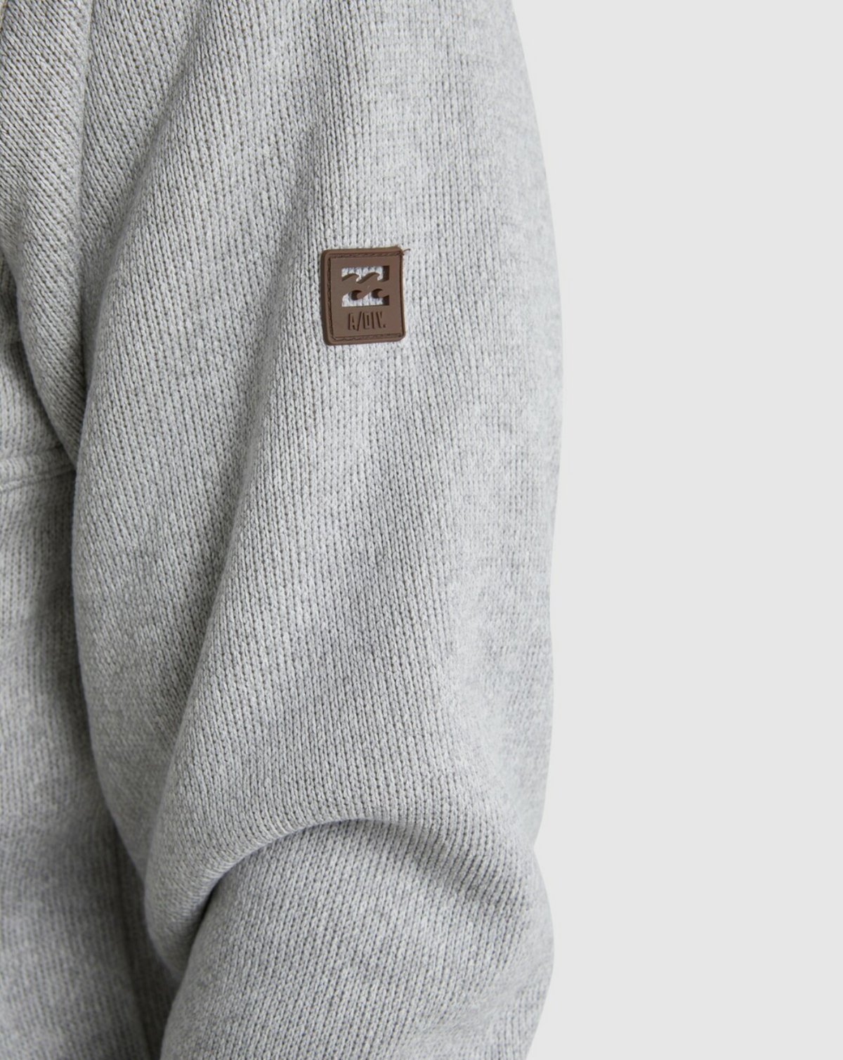 Close-up of the Billabong Boundary Sherpa Zip’s grey knit sleeve with a small brown rectangular patch featuring a logo and "LEVEL" stitched near the upper arm, evoking cozy sherpa lining typical of premium outerwear.
