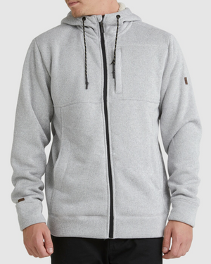 A person wears the Billabong Boundary Sherpa Zip in light gray with black drawstrings, front pockets, and black pants, standing against a plain light background. The hoodie is lined with cozy sherpa for extra warmth.