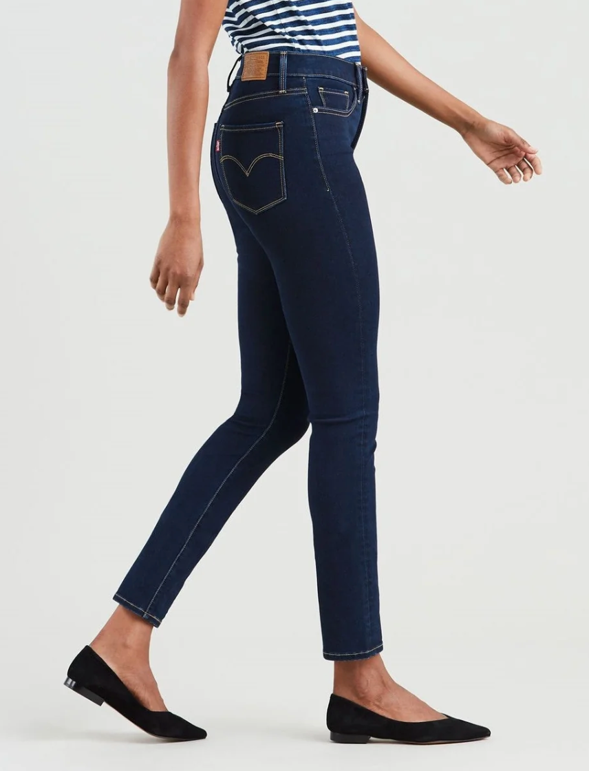 A person walking in dark blue Levi's 312 Shaping Slim Jeans, recognized for their tummy-slimming panel, paired with a striped top and black flats. The image captures a side view from shoulders to feet, with slightly bent arms.
