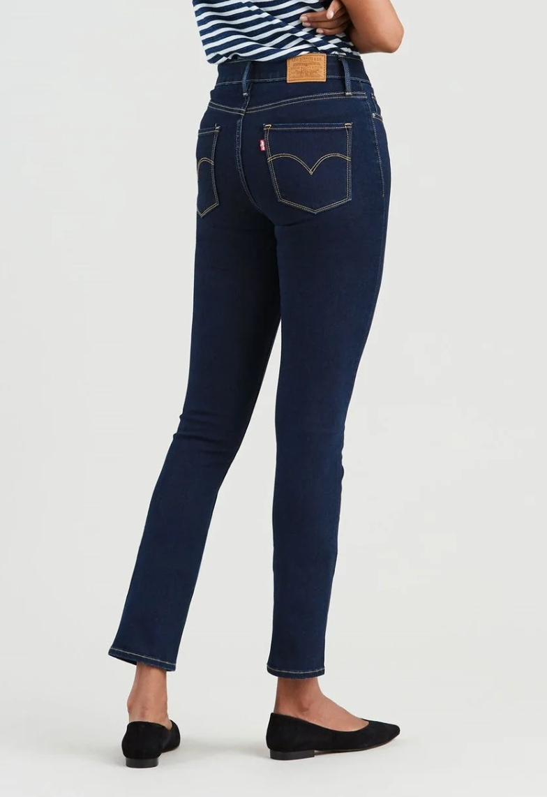 A person wearing Levi's 312 Shaping Slim Jean, featuring visible stitching and a leather patch, stands with crossed arms. The dark blue jeans include a tummy-slimming panel and are paired with a black and white striped top and black flats, as they face away from the camera.