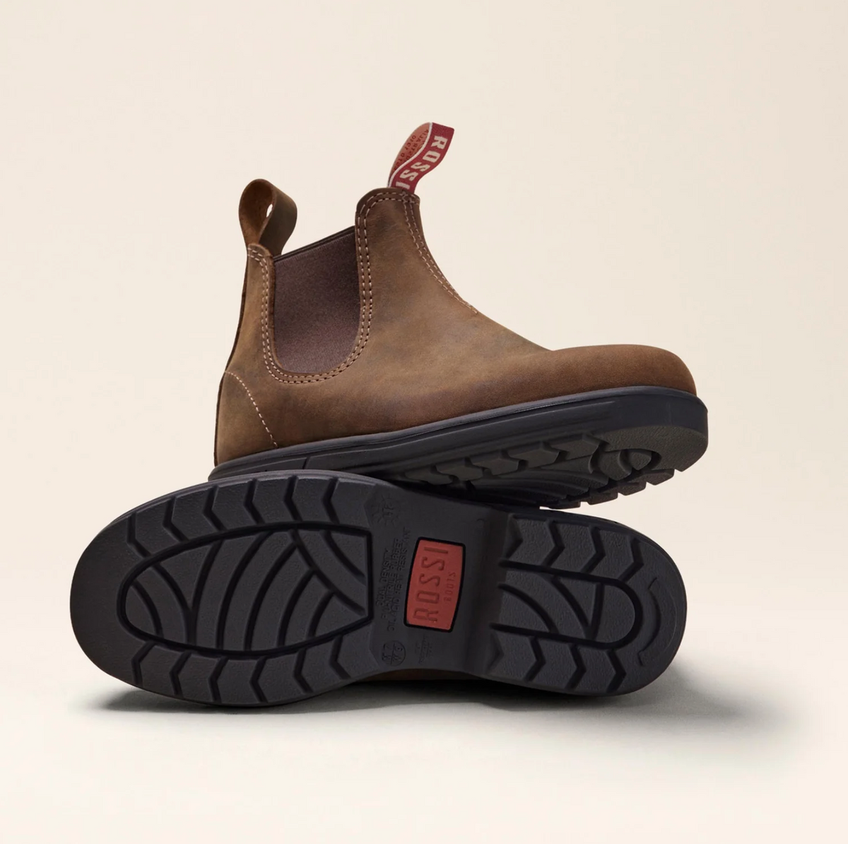 Displayed are the Rossi Endura 303 Elastic Sided Boots in brown full-grain leather with black, slip-resistant soles. One boot stands upright and the other is tilted to show its tread pattern, featuring red pull tabs with "Rossi” text for a stylish and durable look.