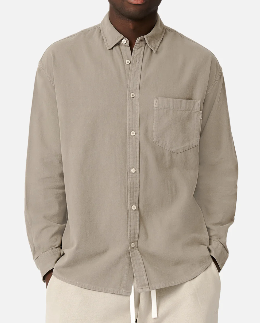 A person in an Industrie The Taylor Shirt, light beige and 100% cotton with a front pocket, stands with hands in pockets. This comfortable button-up shirt is paired with casual white pants against a plain white backdrop.