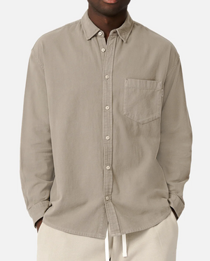 A person in an Industrie The Taylor Shirt, light beige and 100% cotton with a front pocket, stands with hands in pockets. This comfortable button-up shirt is paired with casual white pants against a plain white backdrop.
