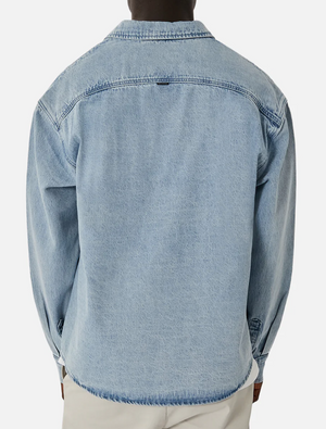 Wearing the Industrie The Lafayette Shirt, a light blue denim button-up with classic collar and long sleeves in 100% cotton, a person stands out against a plain white background. Paired with light-colored pants, the relaxed fit of this stylish piece is captured from behind.