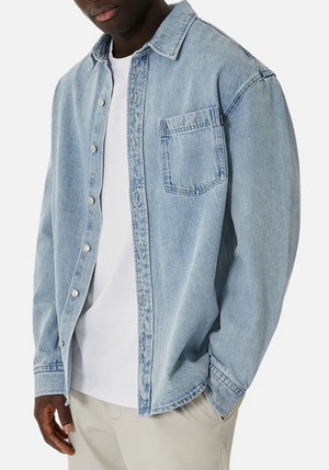 Someone is wearing the Industrie The Lafayette Shirt, a stylish light blue denim button-up over a white t-shirt and beige pants. The 100% cotton, relaxed-fit shirt has a chest pocket and slightly rolled-up sleeves, all set against a plain white background.