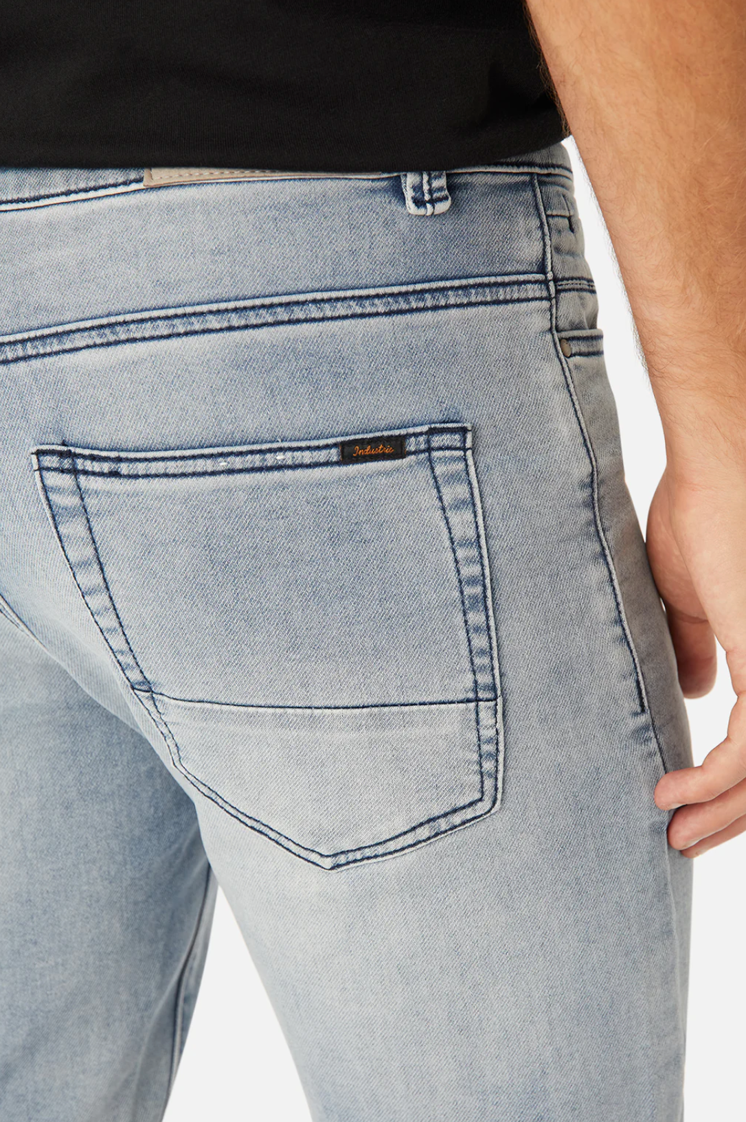 Close-up of the back of a person wearing Industrie The Denim Drifter NC Pant in light blue, skinny fit and a black top. The focus is on the back pocket of the jeans, highlighting the stitching and texture of the stretchy denim. The person is partially turned to the side, with their hand visible by their hip.