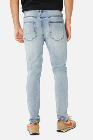 A person wearing Industrie The Denim Drifter NC Pant in light blue and beige Nike sneakers with orange Swooshes is standing with their back turned. The jeans feature back pockets with stitching details. They are also wearing a black shirt.