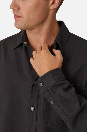 A person wears Industrie The Portland Shirt, a dark gray cotton/linen blend button-up, with the collar slightly turned up. They're touching the collar with their right hand, visible from torso to partial face against a white background.