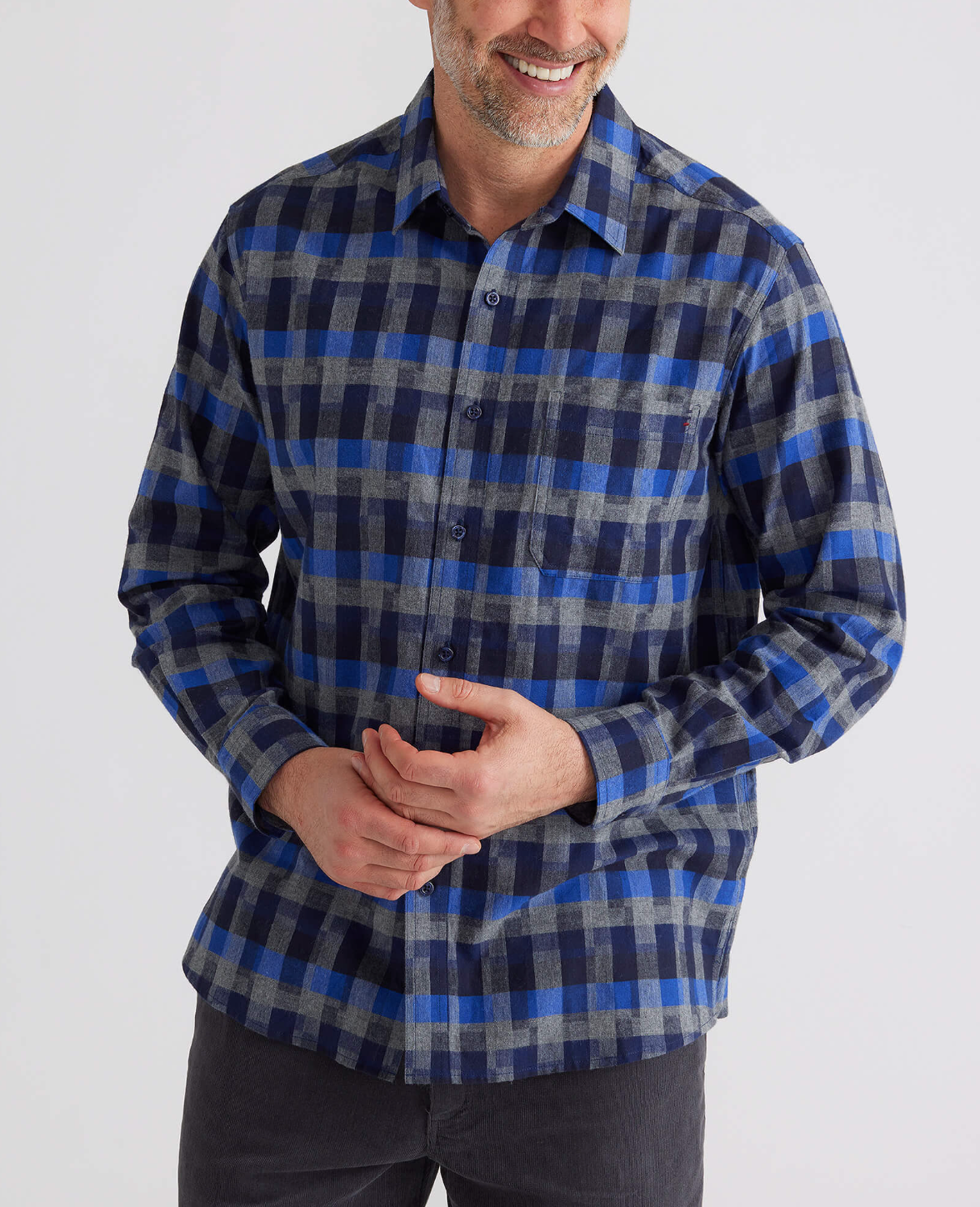 A smiling man with short hair stands against a light background, clasping his hands in front of him. He is wearing the Breakaway Benny Cotton Brush Shirt in a comfortable blue and gray checkered pattern, paired with dark pants.