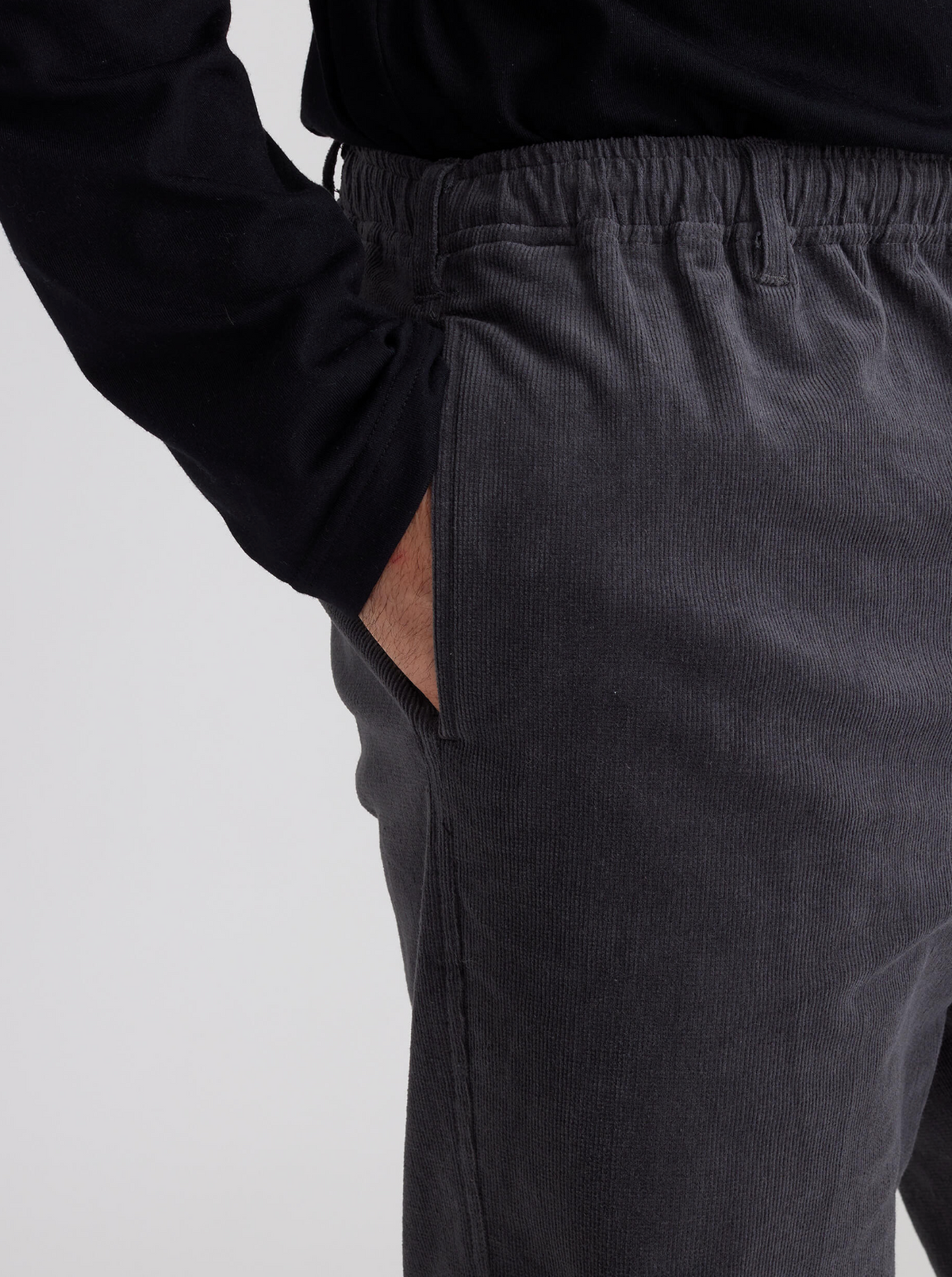 A person wearing a black long-sleeve shirt and Breakaway Cordeaux Pants in gray with an elastic waist, casually resting one hand in the pocket. The cropped photo from shoulder to mid-thigh highlights the fabric's rich texture.