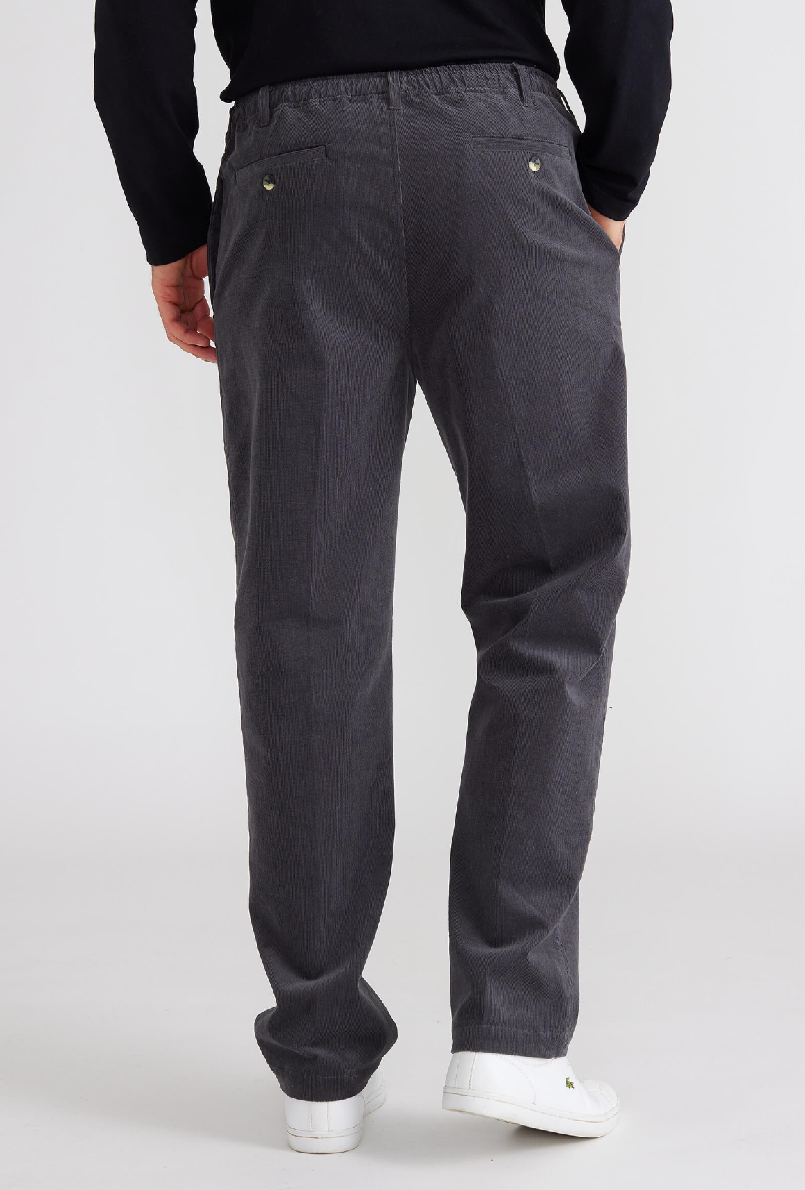 Rear view of someone sporting Breakaway Cordeaux Pant in charcoal gray with an elastic waist and two back button-detail pockets, paired with a black long-sleeved shirt and white sneakers, set against a plain white background.
