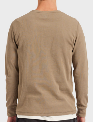 Rear view of a person in an Academy Brand Workers Crew long-sleeve, light brown shirt with rib knit texture, standing against a plain white backdrop. The outfit features dark pants and a comfy crew neck undershirt visible through their short hair.
