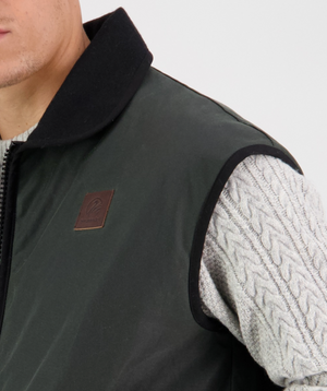 Someone is wearing a dark green Swanndri Foxton Vest Wool Lined with a leather patch, paired with a light grey cable-knit sweater underneath. The image is cropped to show the upper torso and part of the face against a plain background.