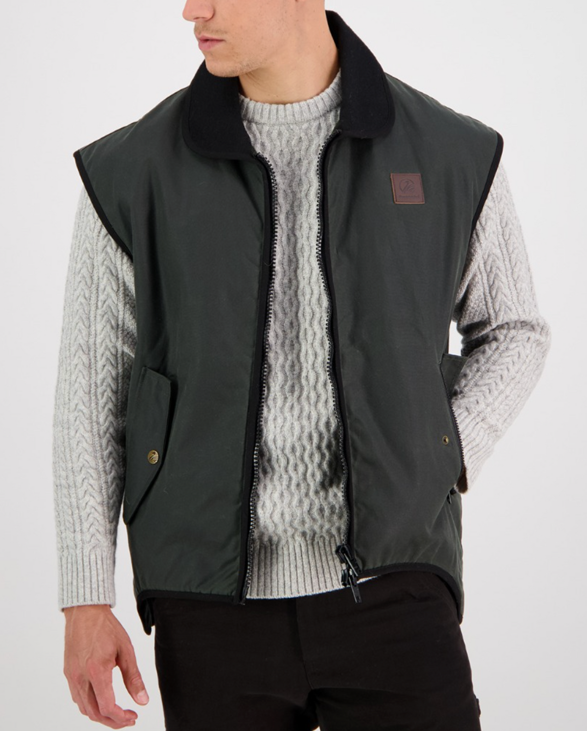 A person is posed against a white background, wearing a Swanndri Foxton Vest Wool Lined in dark green. The waterproof, sleeveless jacket features a black collar, zipper front, and two pockets. They have layered it over a light gray cable-knit sweater paired with black pants and are gazing to the side.