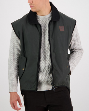 A person is posed against a white background, wearing a Swanndri Foxton Vest Wool Lined in dark green. The waterproof, sleeveless jacket features a black collar, zipper front, and two pockets. They have layered it over a light gray cable-knit sweater paired with black pants and are gazing to the side.