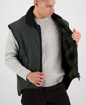 A person wearing a gray cable-knit sweater and a Swanndri Foxton Vest Wool Lined jacket with a black collar holds the jacket open to show its green plaid inner lining, looking down with a neutral expression.