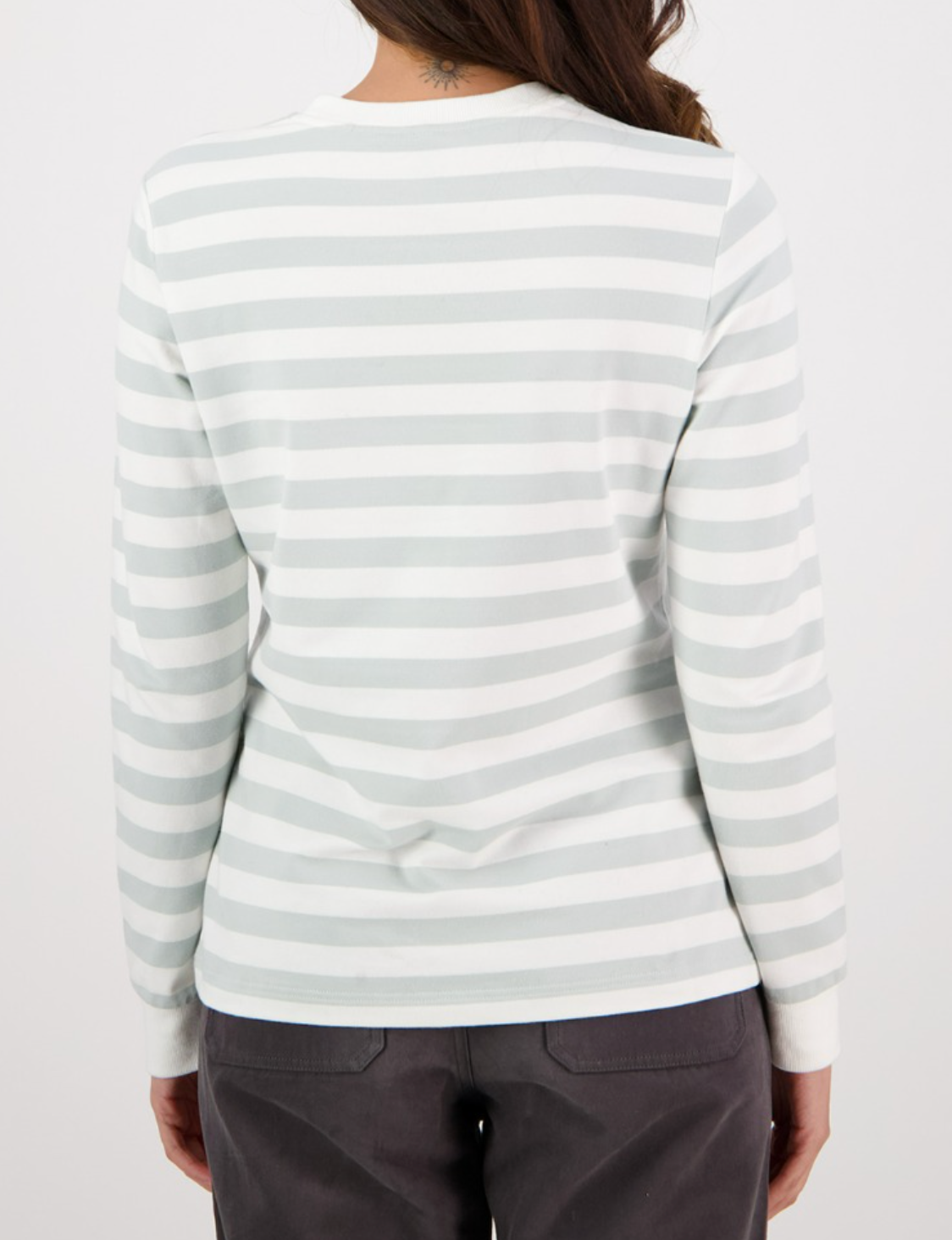 The image shows a person wearing a relaxed-fit Swanndri Papamoa Stripe Long Sleeve Tee in light gray and white stripes. Their long, dark hair falls down their back as they pair it with dark pants, set against a clean white backdrop.