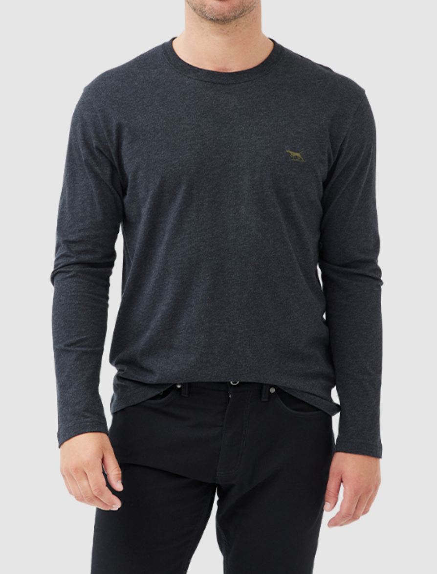 A person is wearing a Rodd & Gunn Long Sleeve Gunn T-Shirt, dark gray and 100% cotton, with a small embroidered logo on the left chest. The shirt has a sports fit and is paired with black pants against a light gray background. The person's face is not visible.