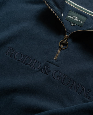 The close-up highlights the Rodd & Gunn Alford Forest Sweat, a navy mid-weight jersey pullover with a half zip. "RODD & GUNN" is subtly embroidered in matching thread, and the metal circular zipper pull reveals the tag with the brand's distinctive white and green logo.