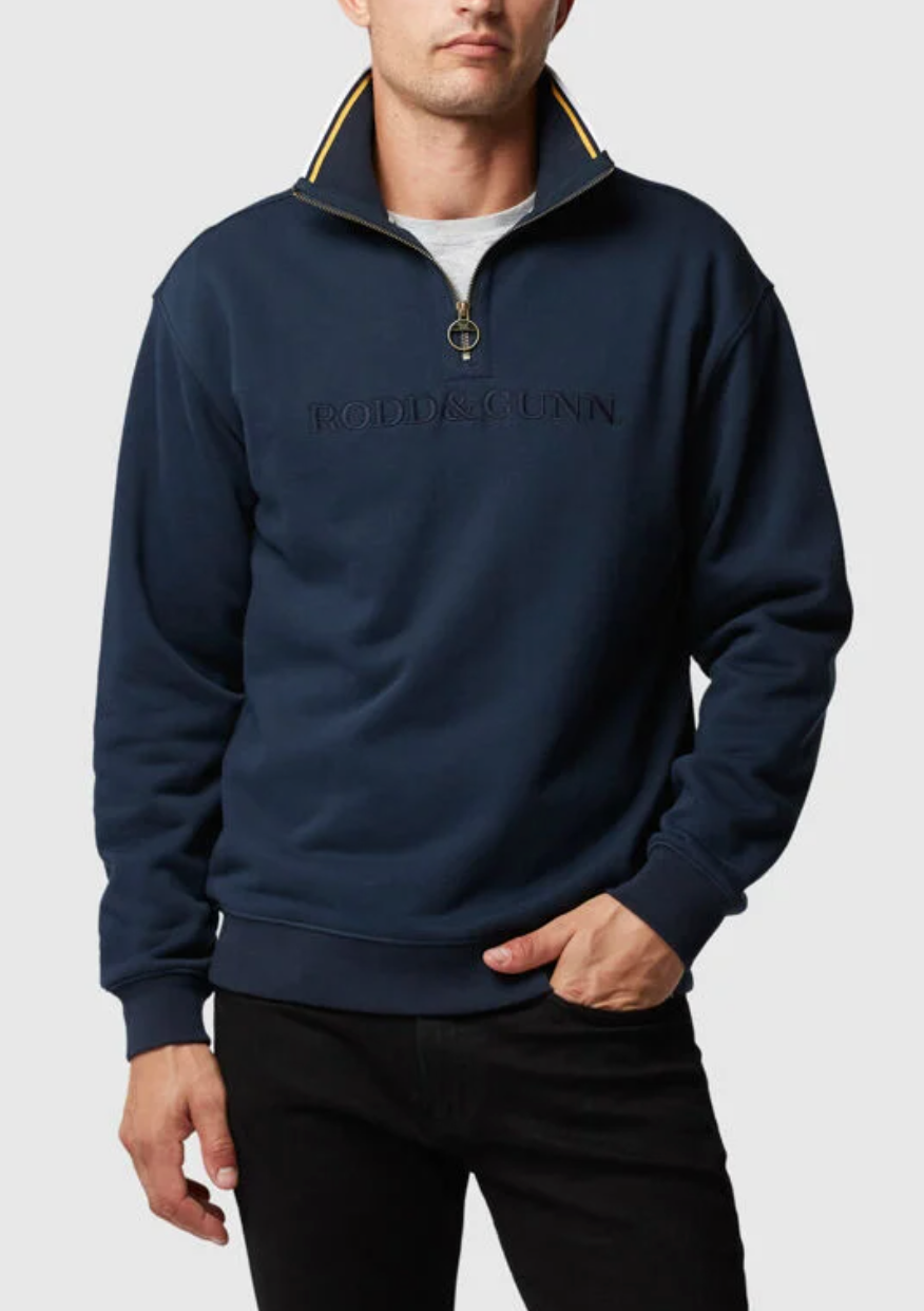 Dressed in a navy mid-weight Rodd & Gunn Alford Forest Sweat with "Rodd & Gunn" emblazoned on the front, a person exudes style with its contrasting half zip and collar, as their hand slips into the pocket of black pants against a simple light gray backdrop.