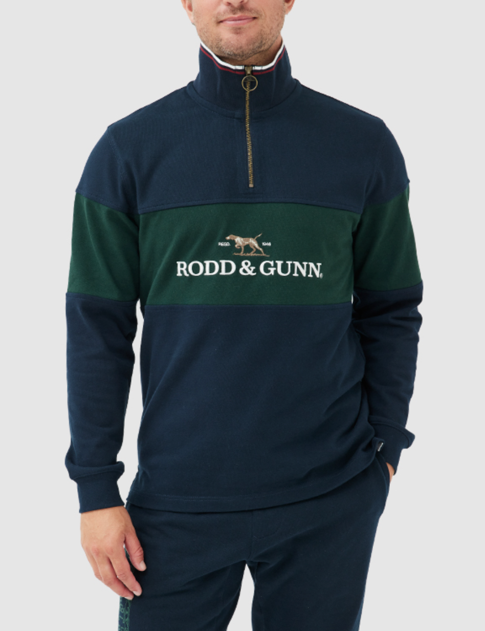 Man wearing a Rodd & Gunn Foresters Peak Sweat, a navy and green quarter-zip pullover made from 100% cotton mid-weight jersey. It features "Rodd & Gunn" on the chest, an embroidered dog logo, funnel neck, and zip detail. He pairs it with matching dark pants.