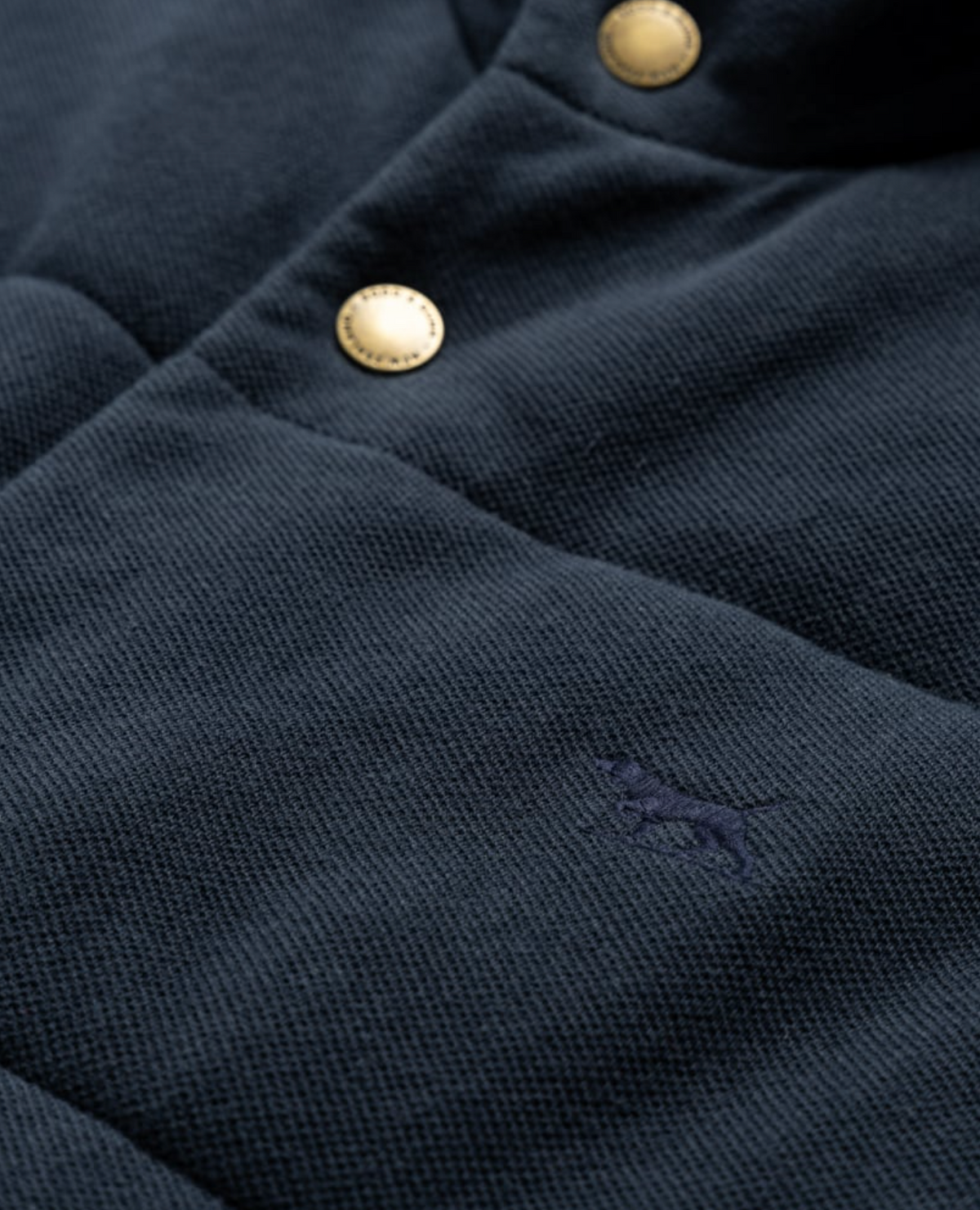 Close-up of the Rodd & Gunn Lake Ferry Vest, a textured dark fabric vest with gold buttons, a small embroidered dog logo in dark blue, crafted from USA Cotton for lightweight insulation.