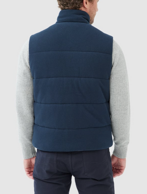 A person facing away is wearing the Rodd & Gunn Lake Ferry Vest in navy blue, featuring lightweight insulation, over a light gray USA Cotton sweater. They have short hair, dark pants, and stand against a plain, light-colored background.