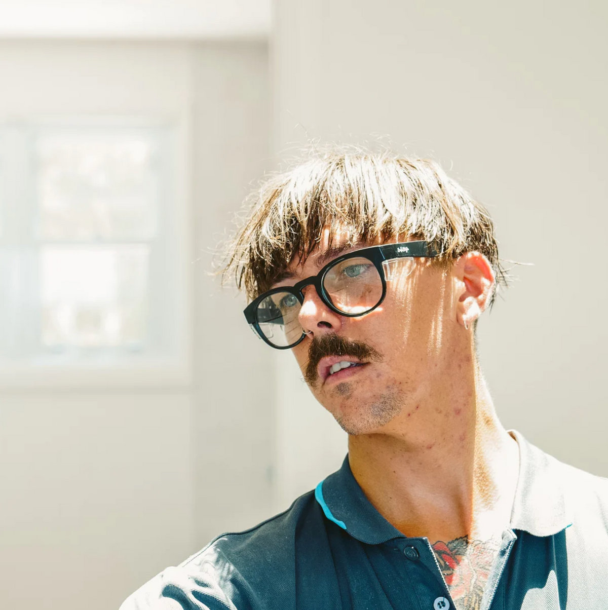 In a brightly lit room, a person with short-cropped hair and glasses featuring SafeStyle Cruisers Black Frame Clear Lens glances sideways. They wear a dark shirt that reveals tattoos on their neck. A window provides UV protection while allowing natural light to enter.