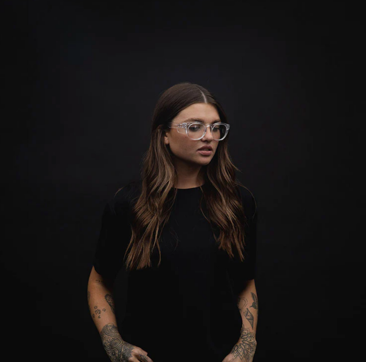 A woman with long brown hair, adorned with tattoos and wearing SafeStyle Cruisers Clear Frame Clear Lens glasses, stands against a dark background. Sporting a black shirt, she exudes both style and safety as she gazes to the side with a neutral expression.