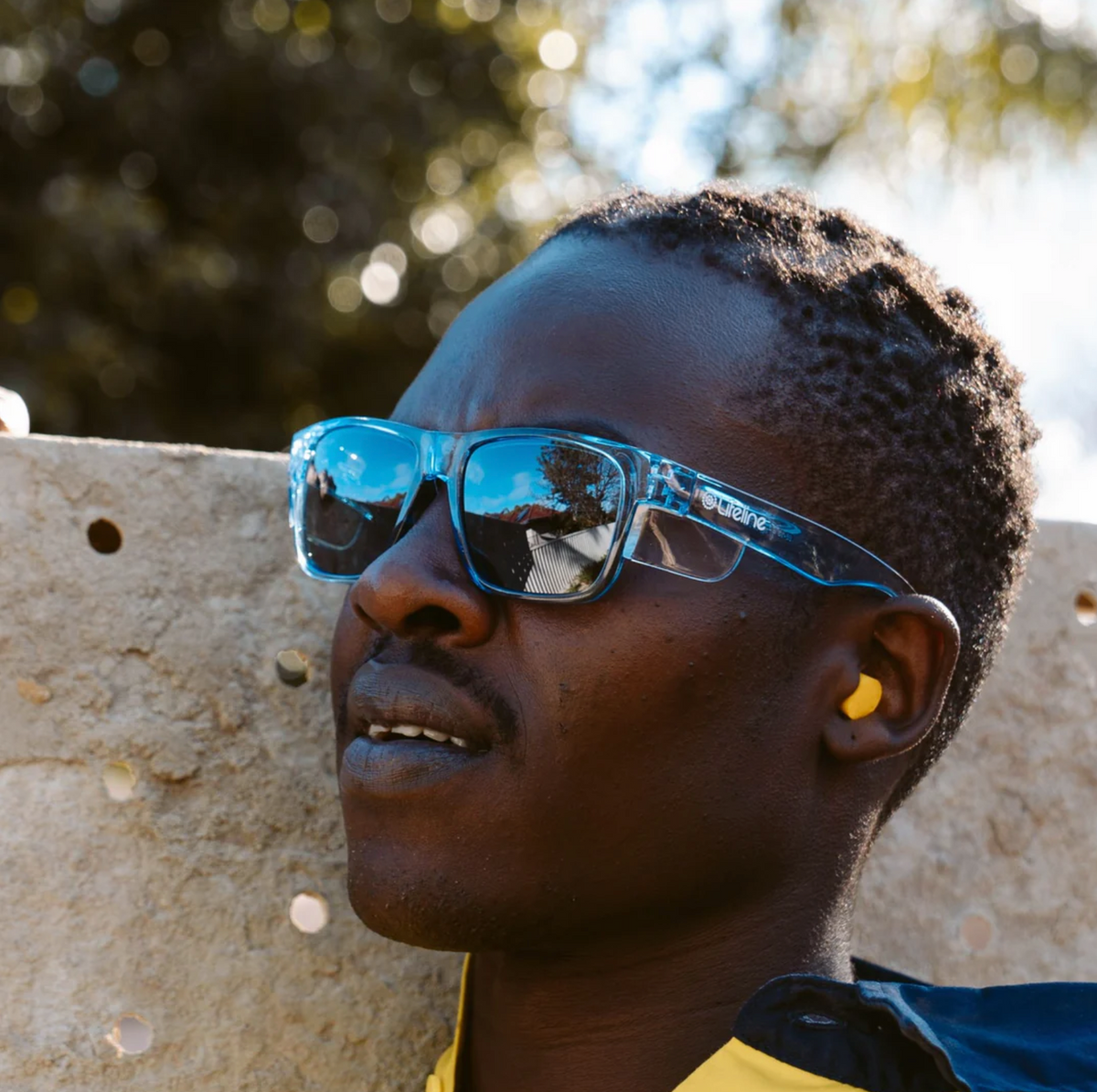 Wearing SafeStyle Fusions sunglasses with blue frames and polarised lenses, a person in yellow earplugs gazes up outside. The UV-protected lenses reflect natural scenery, while trees and the sky blur in the background.