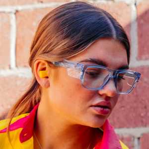A person with long brown hair wears SafeStyle Fusions blue-frame clear lens glasses and a colorful shirt with a red collar. They have a yellow earplug in one ear. The background is a textured brick wall.