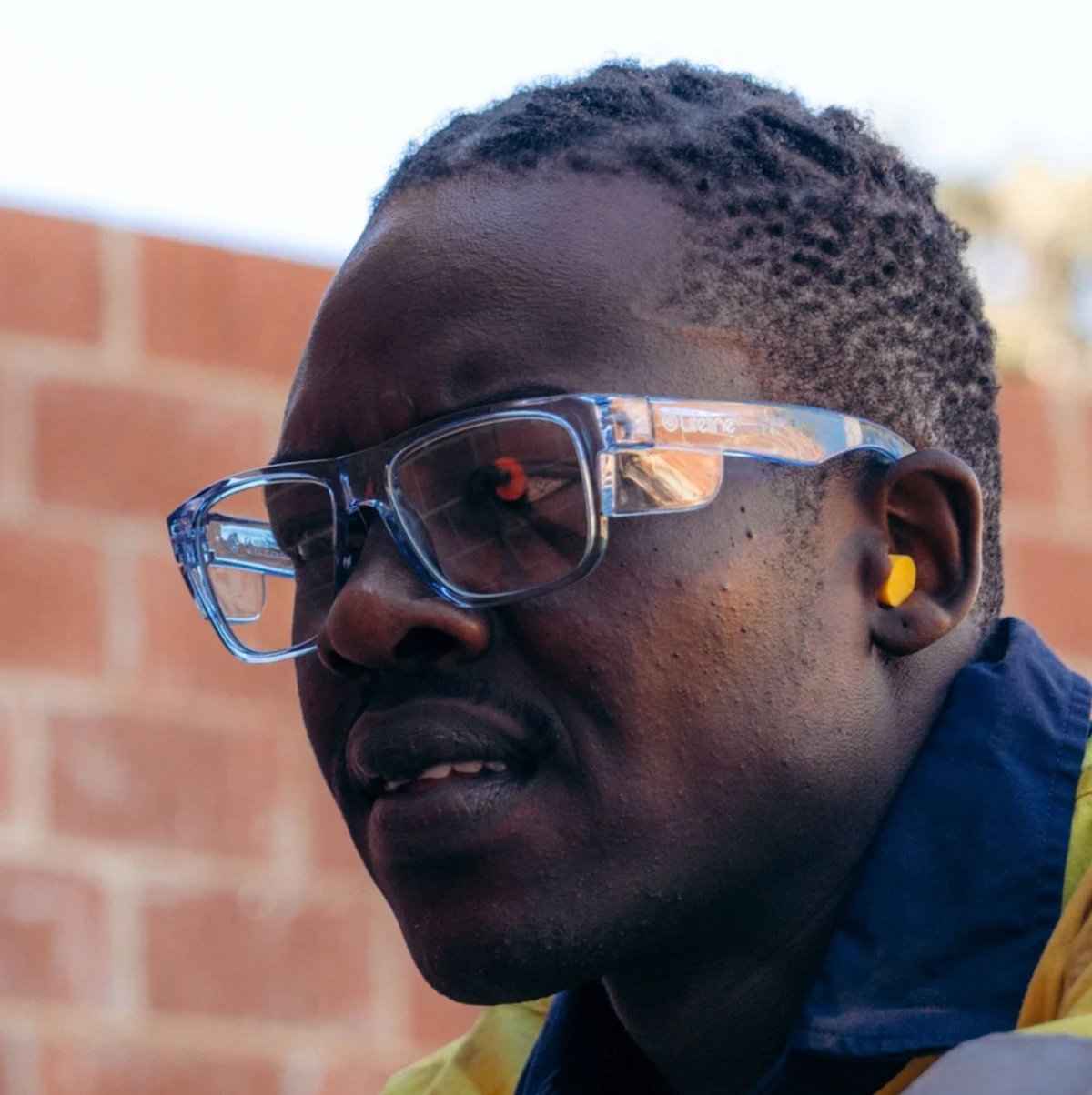 A person wearing SafeStyle Fusions safety glasses with a blue frame and clear lens, offering UV protection and high safety ratings, gazes intently at something off-camera. With short hair, they're in front of a brick wall, wearing a yellow and navy collared shirt.