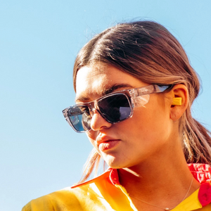 A person with long brown hair, wearing SafeStyle Fusions Graphite Frame Tinted Lens sunglasses and a yellow shirt, looks to the side against a clear blue sky. They are also wearing a yellow earplug in one ear.