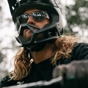 A close-up of a person with long hair wearing SafeStyle Fusions Graphite Frame Tinted Lens sunglasses and a black full-face helmet, likely for biking or motorcycling. The background is blurred, indicating an outdoor setting with trees. The person appears ready for an adventurous ride, benefiting from added UV protection.