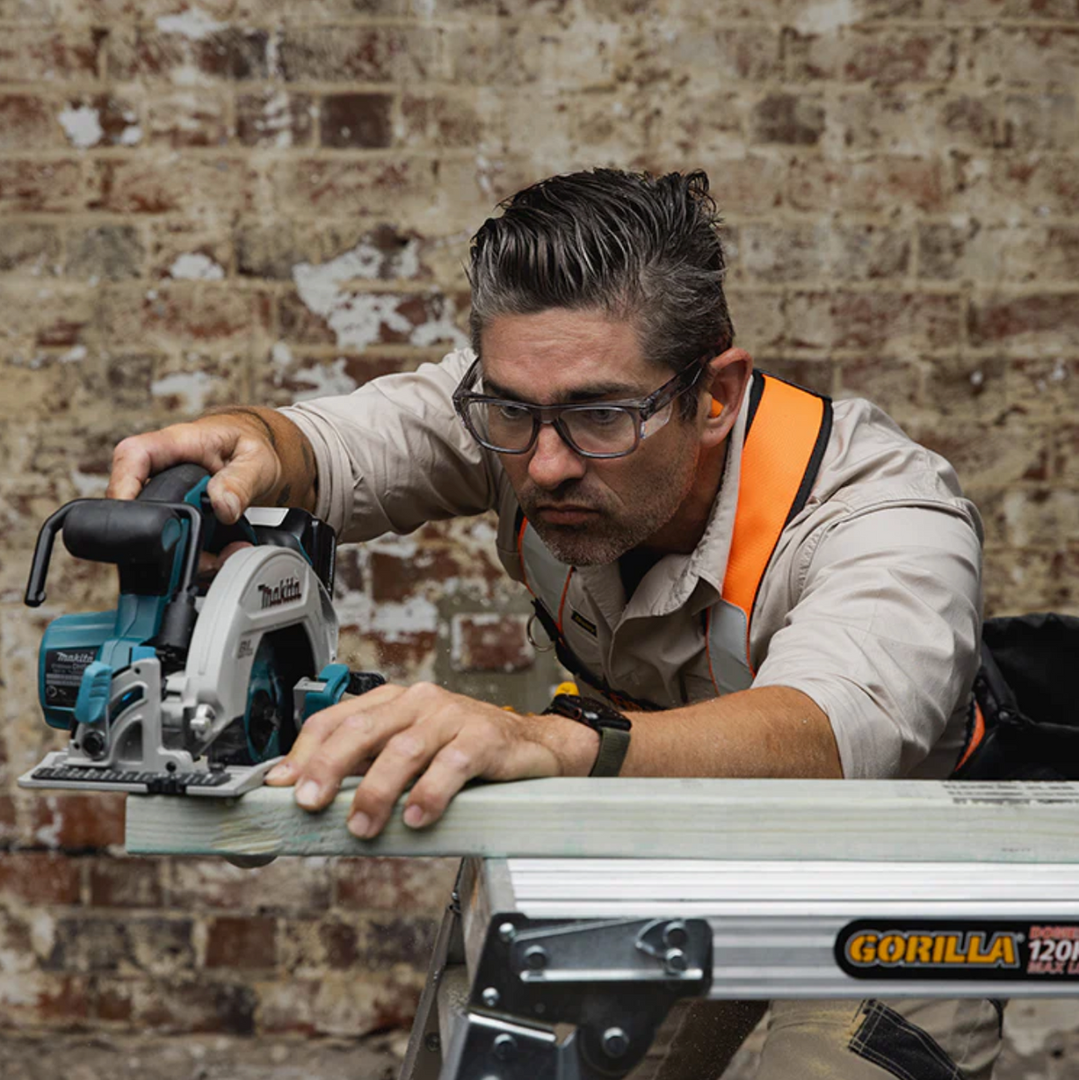 A man wearing SafeStyle Fusions Graphite Frame Clear Lens safety glasses and an orange harness uses a cordless circular saw to cut a wooden plank on a metal sawhorse. He is focused, framed by the brick wall in the background, ensuring precision and protection.