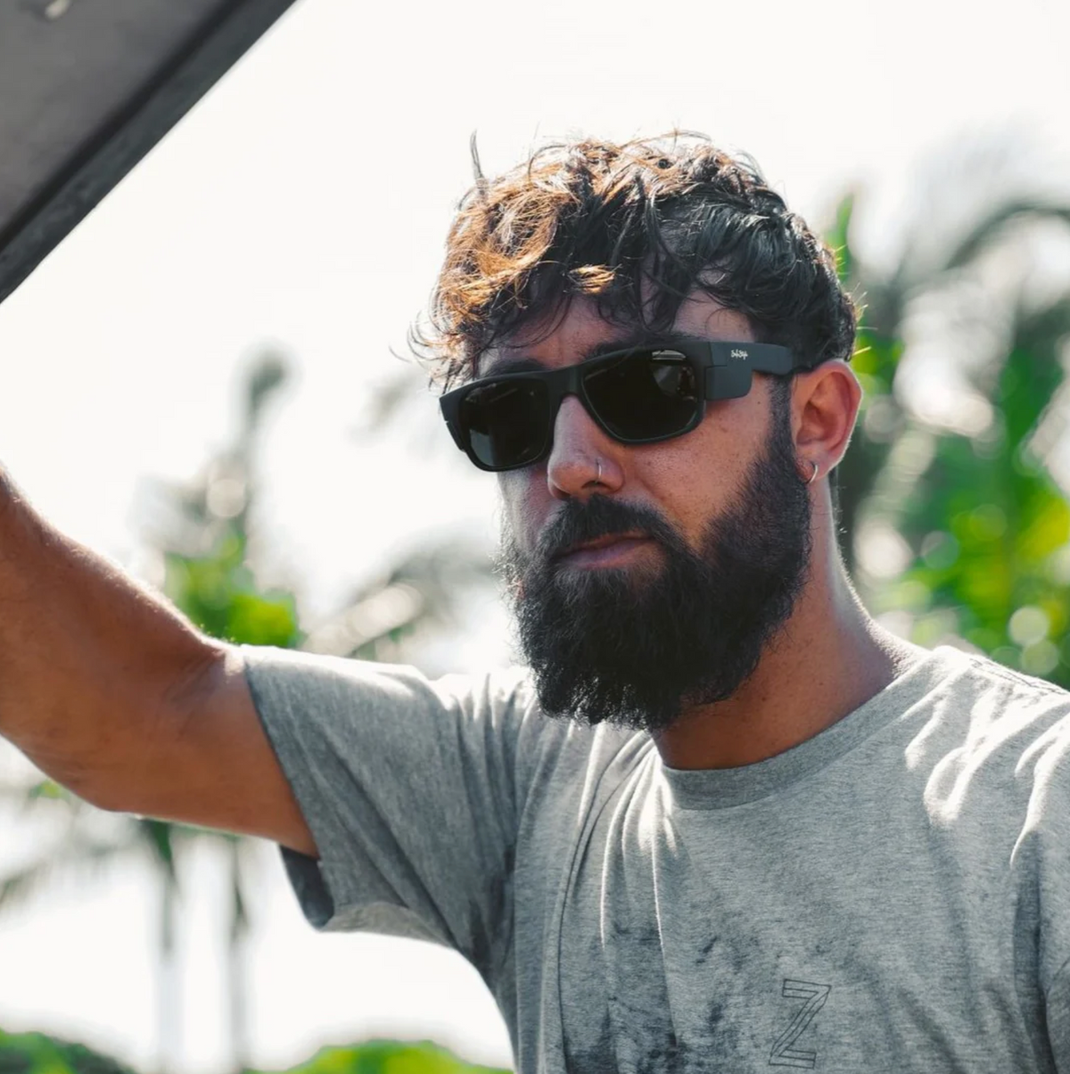 A bearded man wearing SafeStyle Fusions Matte Black sunglasses stands outdoors in a gray t-shirt, gripping a ledge. Sunlight highlights his hair as he looks through the polarized lenses, with palm trees visible in the blurred background.