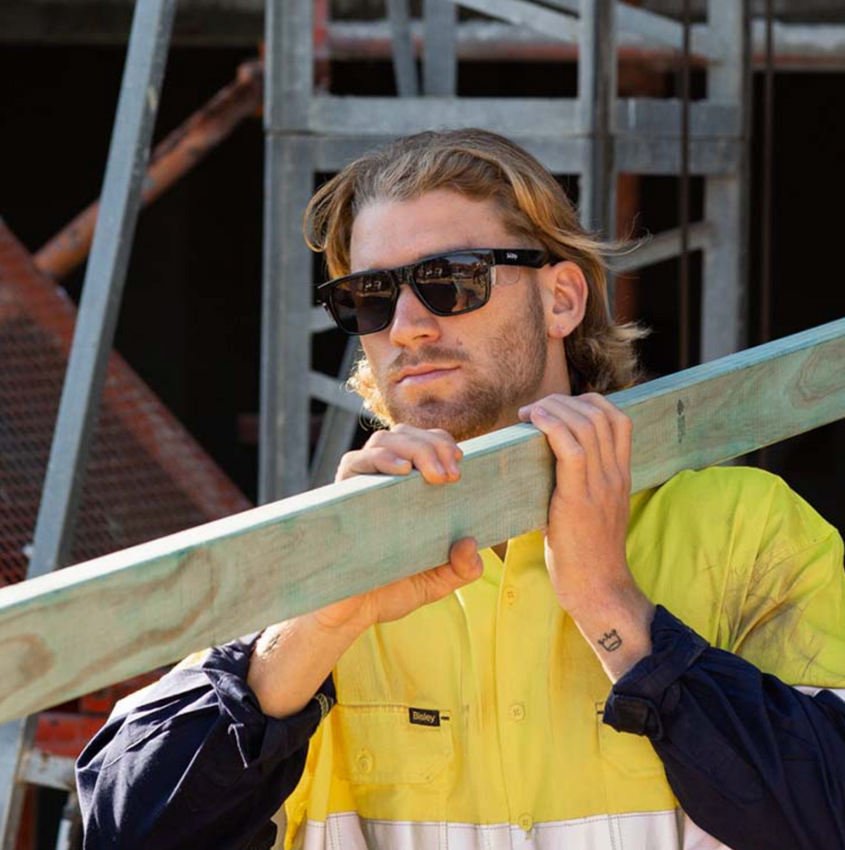 Wearing SafeStyle Fusions Black Frame Polarised Lens sunglasses and a high-visibility yellow and navy work shirt, a person shoulders a wooden plank at a construction site with scaffolding in the background.