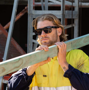 Wearing SafeStyle Fusions Black Frame Polarised Lens sunglasses and a high-visibility yellow and navy work shirt, a person shoulders a wooden plank at a construction site with scaffolding in the background.