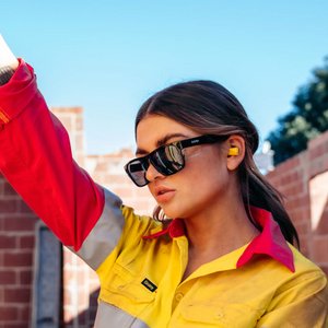 Wearing a high-visibility work shirt, a person stands confidently outside against a brick structure. With ear protection and raised arm under the clear blue sky, they wear SafeStyle Fusions Black Frame Tinted Lens sunglasses for added UV protection and safety.