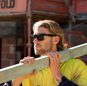 A person with long hair wears SafeStyle Fusions sunglasses, featuring a black frame and tinted lenses with UV protection. They stand at a construction site in a yellow work shirt, carrying a wooden beam on their shoulder, glancing to the side amidst scaffolding and bricks.