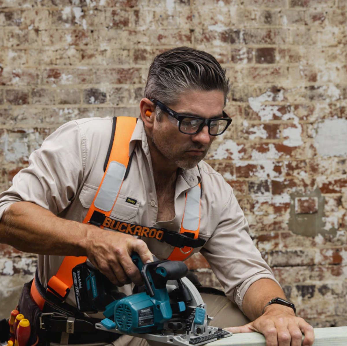 A man wearing SafeStyle Fusions Black Frame Clear Lens safety glasses and an orange harness uses a power saw to cut a plank of wood in front of a worn brick wall.