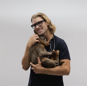 A person with SafeStyle Fusions black frame clear lens glasses and long hair smiles while holding a small brown dog. They wear a dark outfit with a stethoscope, standing against a plain white background.