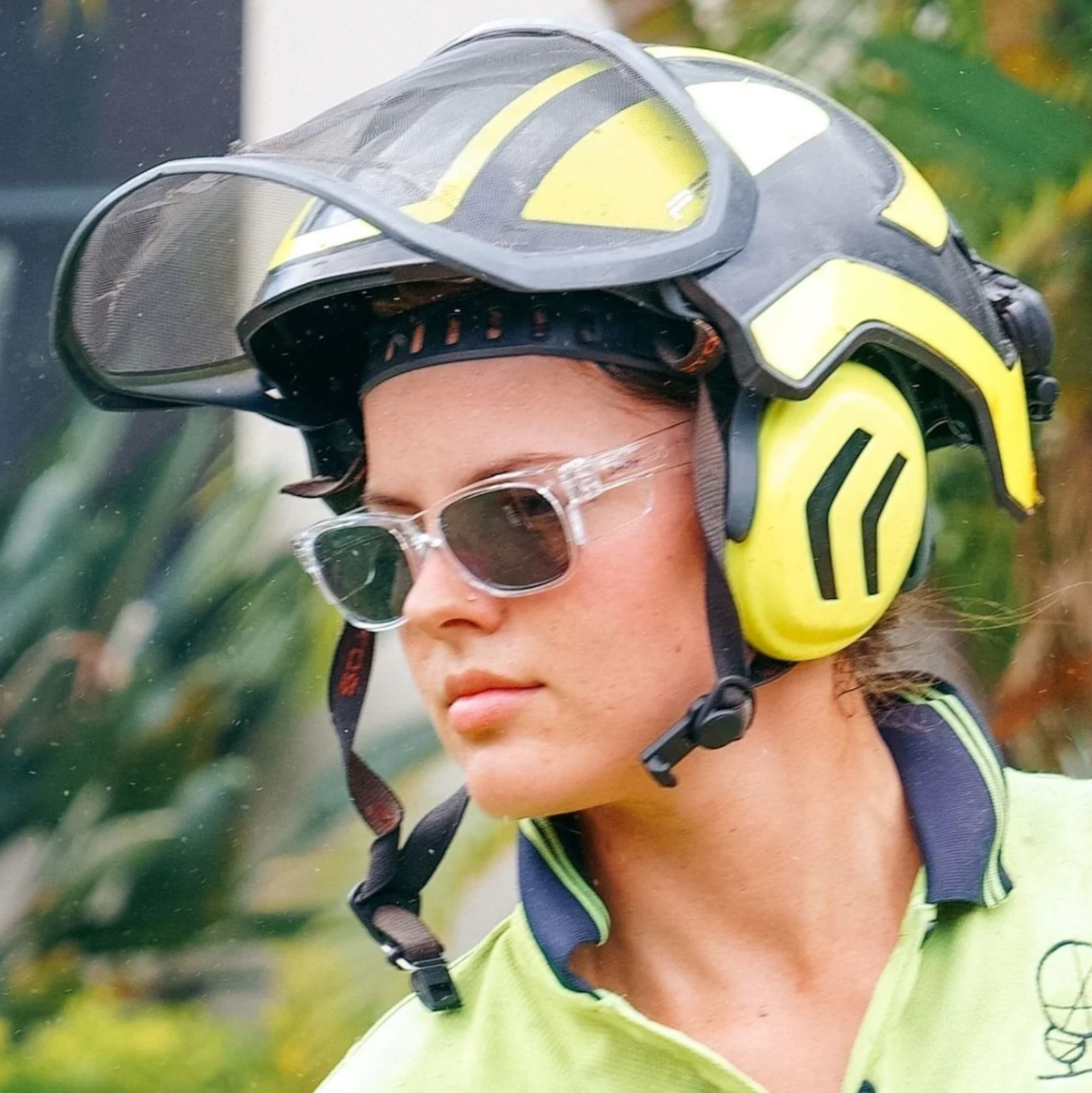 A person wearing SafeStyle Fusions Clear Frame Polarised Lens safety sunglasses, a green and black protective helmet with a visor and ear protection, and a green shirt is outdoors with blurred greenery in the background, enjoying effective UV protection.